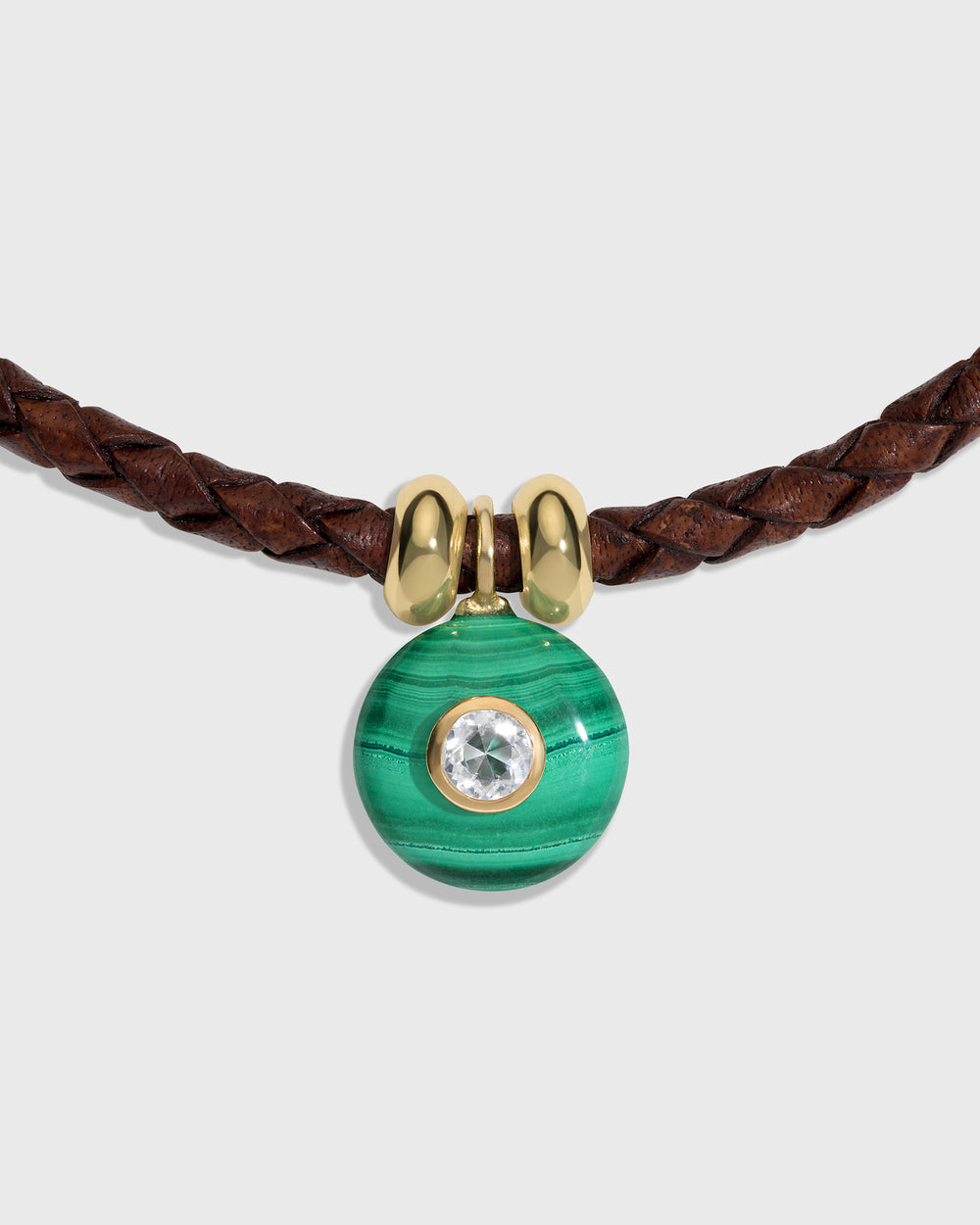 A close-up of a necklace featuring a brown braided leather cord, a polished gold bow-shaped connector, and a round green malachite pendant with a clear crystal in the center, set against a light gray background.