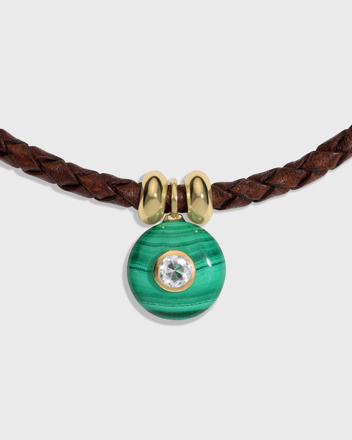 A close-up of a necklace featuring a brown braided leather cord, a polished gold bow-shaped connector, and a round green malachite pendant with a clear crystal in the center, set against a light gray background.
