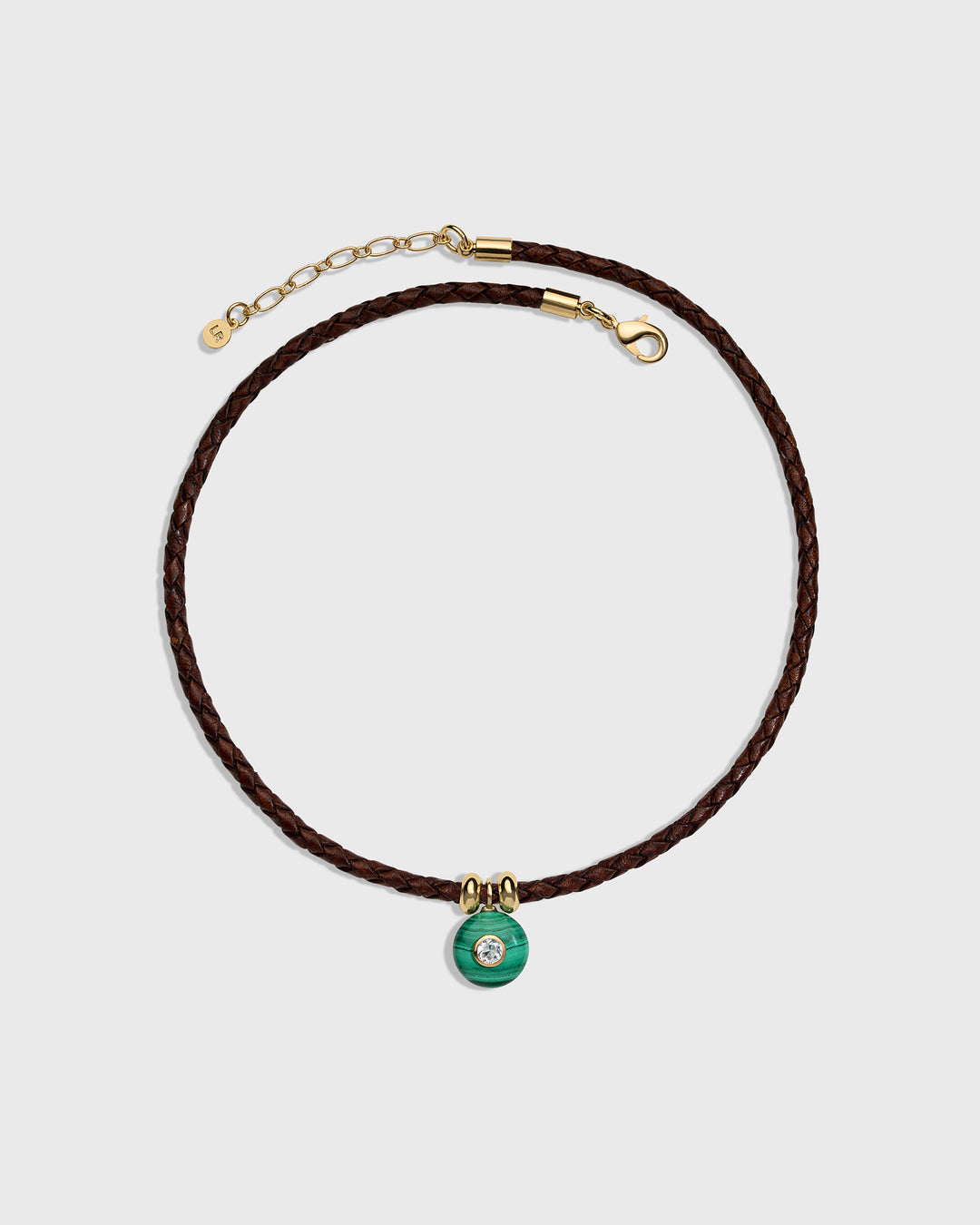 A dark brown braided cord necklace with a small green circular charm featuring a clear central stone, accented with gold-tone hardware, shown on a light gray background.