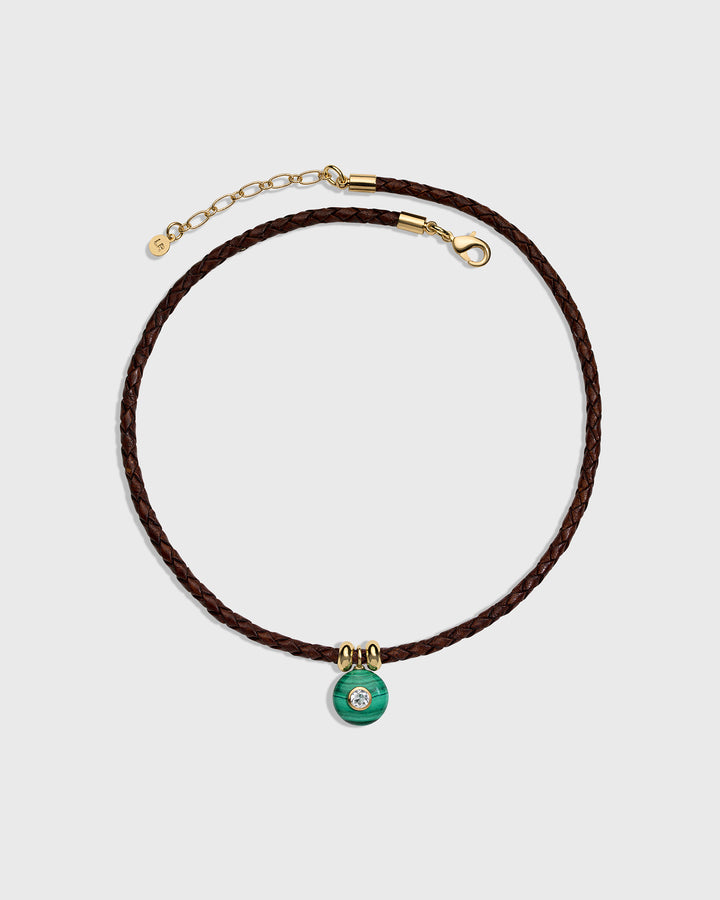 A dark brown braided cord necklace with a small green circular charm featuring a clear central stone, accented with gold-tone hardware, shown on a light gray background.
