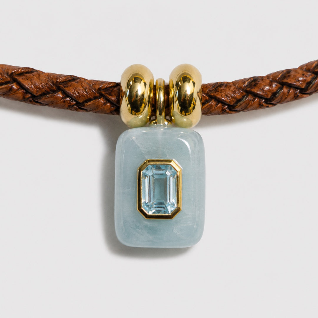 Jewelry with a aquamarine stone on a braided brown cord against a light grey background