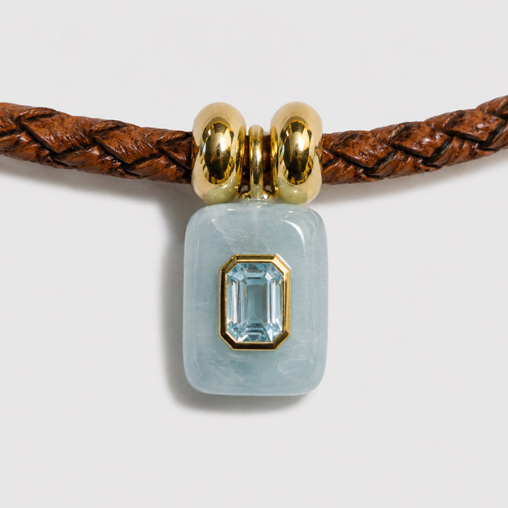 Jewelry with a aquamarine stone on a braided brown cord against a light grey background