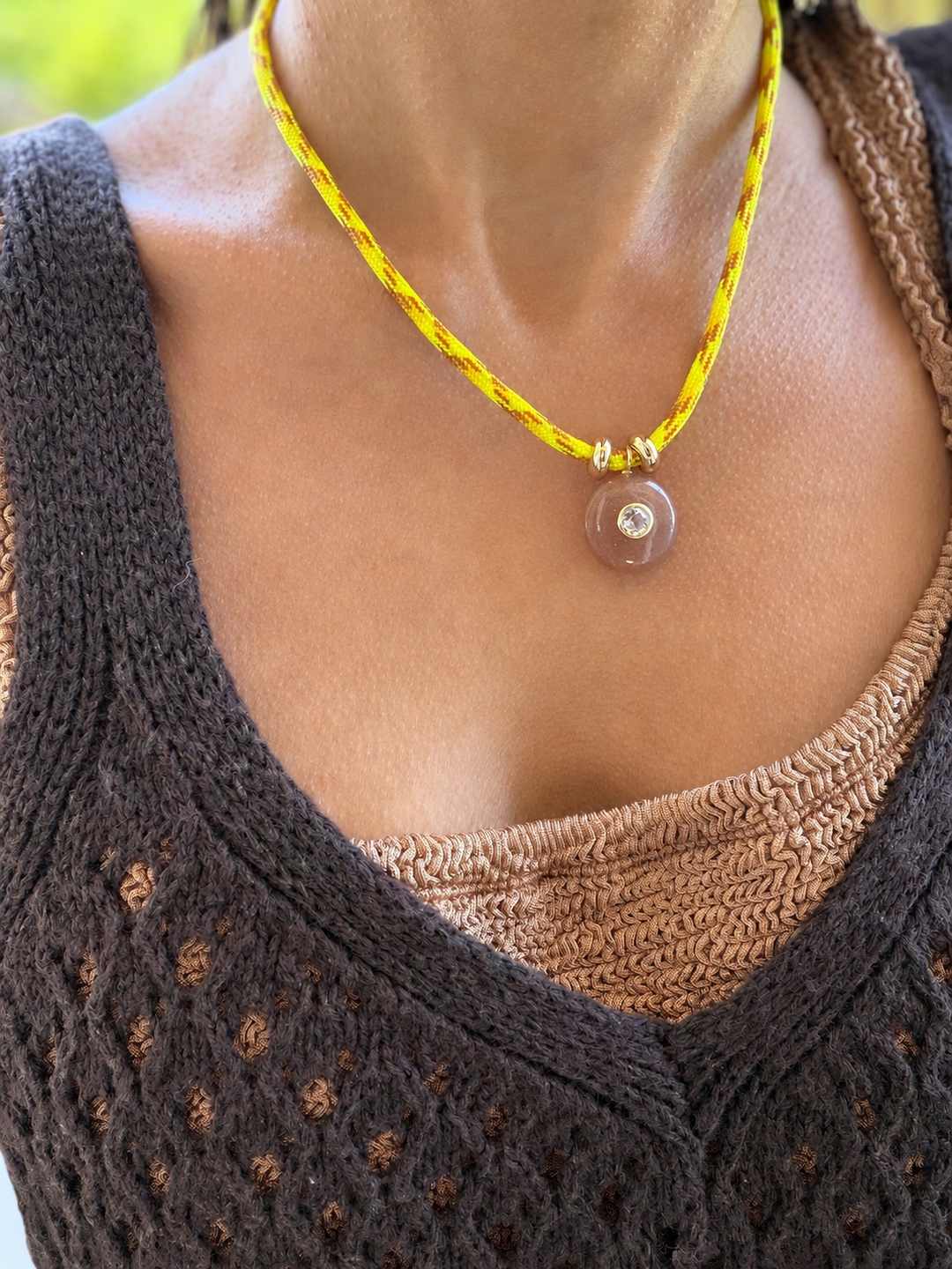 Necklace with a yellow cord and pink pendant worn by a person in a textured brown top.