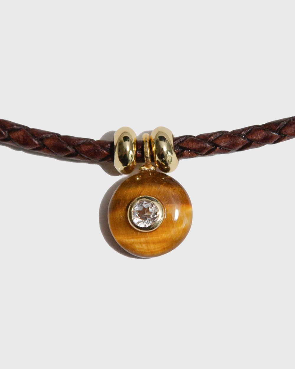 Brown braided leather bracelet with a gold clasp and a round amber bead with a clear stone on a light gray background.